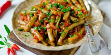 su tavolo apparecchiato tagliere con sopra un piatto con fagiolini in umido con pomodorini e prezzemolo e una forchetta sulla destra peperoncini