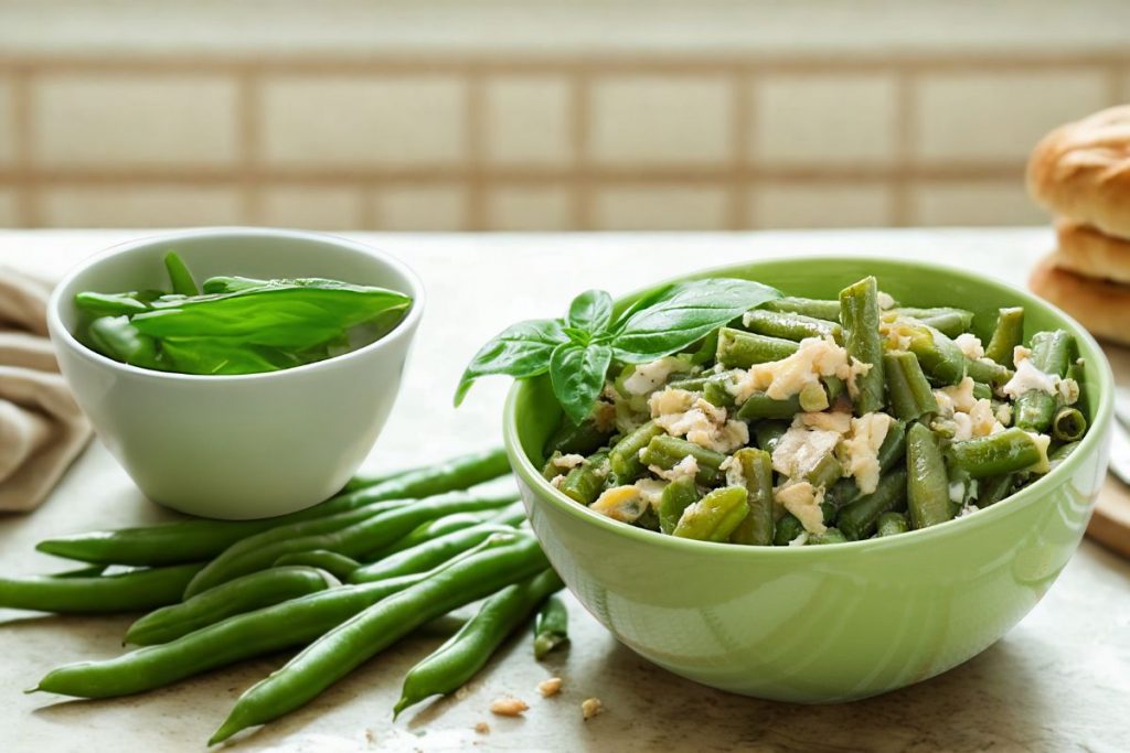Fagiolini lessi: tempo di cottura e come condirli 5 su tavolo da cucina, ciotola verde con fagiolini lessi e parmigiano, accanto un'altra ciottola con basilico e a destra fette di pizza