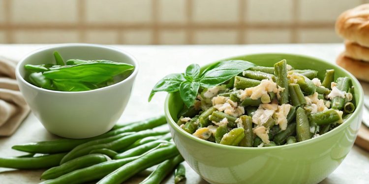 su tavolo da cucina, ciotola verde con fagiolini lessi e parmigiano, accanto un'altra ciottola con basilico e a destra fette di pizza