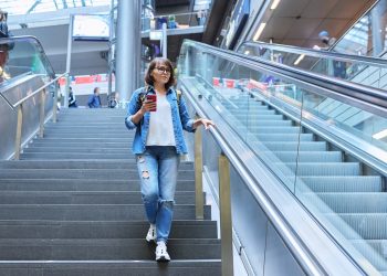 donna con cellulare in mano vestita con camicia di jeans e jeans scende le scale dell'aeroporto per aumentare il suo Neat