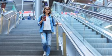 donna con cellulare in mano vestita con camicia di jeans e jeans scende le scale dell'aeroporto per aumentare il suo Neat