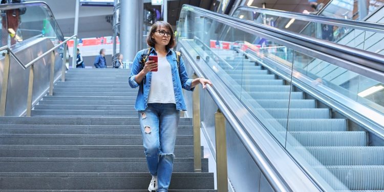 donna con cellulare in mano vestita con camicia di jeans e jeans scende le scale dell'aeroporto per aumentare il suo Neat
