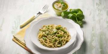 su tavolo bianco apparecchiato piatto bianco con pasta con fagiolini dietro basilico e accanto forchetta gialla e tovagliolo giallo