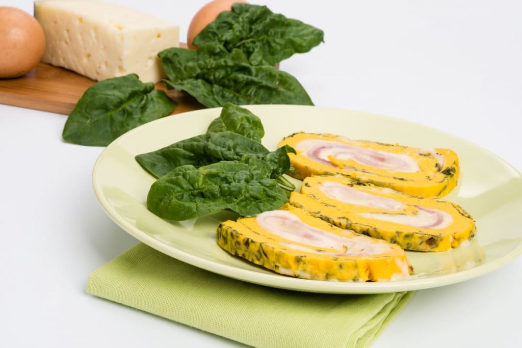 in primo piano un piatto verde chiaro con rotolo di frittata decorato con una foglia di spinacino