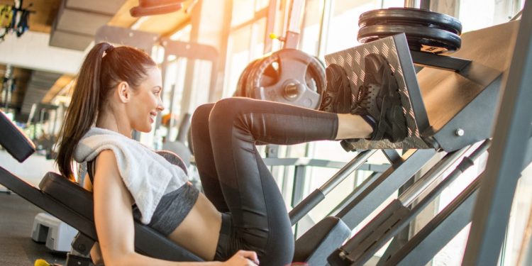donna in palestra con asciugamano sulle spalle esegue con scheda palestra esercizio con la leg press per tonificare le gambe