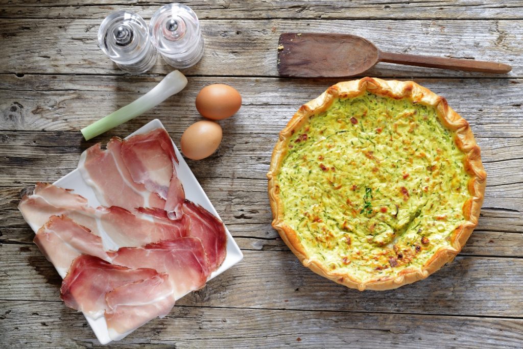 torta salata zucchina e speck su una tavola di legno, sulla sinistra piatto bianco con fette di speck e accanto 2 uova e un pezzo di porro, saliera e pepiera e dietro un cucchiaio di legno