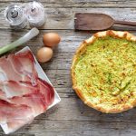 torta salata zucchina e speck su una tavola di legno, sulla sinistra piatto bianco con fette di speck e accanto 2 uova e un pezzo di porro, saliera e pepiera e dietro un cucchiaio di legno