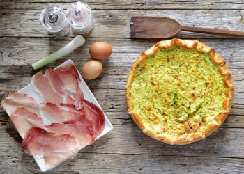 torta salata zucchina e speck su una tavola di legno, sulla sinistra piatto bianco con fette di speck e accanto 2 uova e un pezzo di porro, saliera e pepiera e dietro un cucchiaio di legno