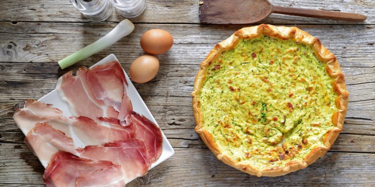 torta salata zucchina e speck su una tavola di legno, sulla sinistra piatto bianco con fette di speck e accanto 2 uova e un pezzo di porro, saliera e pepiera e dietro un cucchiaio di legno