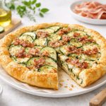 Torta salata zucchina e speck: la ricetta facile senza pasta sfoglia 1 In primo piano su un tavolo da cucina chiaro c'è una torta salata con zucchine e speck. Sulla destra un coltello mentre sullo sfondo ci sono delle zucchine su un tagliere in legno e delle fettine di speck su un piattino.