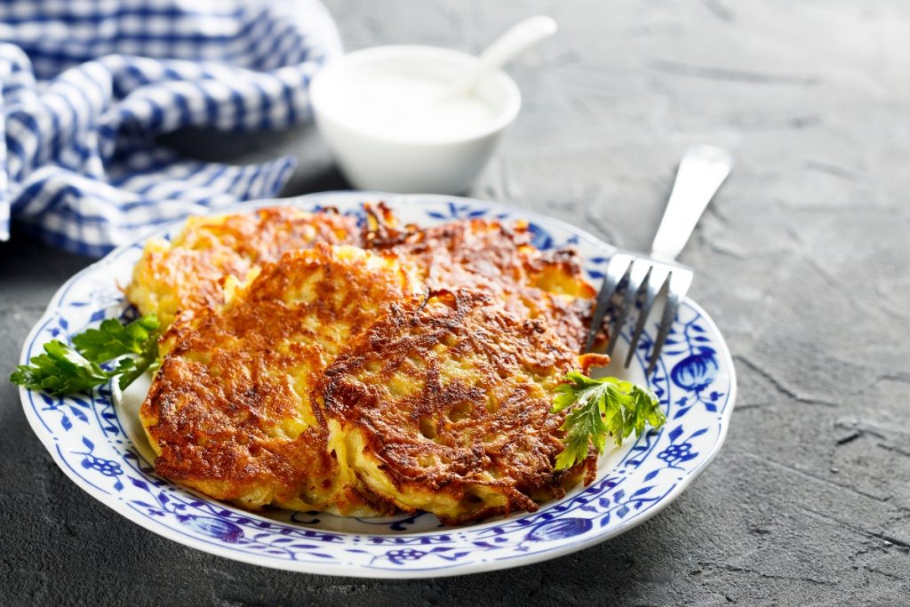 Su tavolo di marmo, piatto bianco con fiori blu con rosti di patate e sotto insalata verde, sul bordo del piatto 1 forchetta e dietro ciotola piccolo con salsa di yogurt e un canovaccio