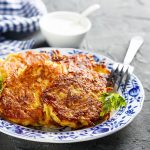 Su tavolo di marmo, piatto bianco con fiori blu con rosti di patate e sotto insalata verde, sul bordo del piatto 1 forchetta e dietro ciotola piccolo con salsa di yogurt e un canovaccio