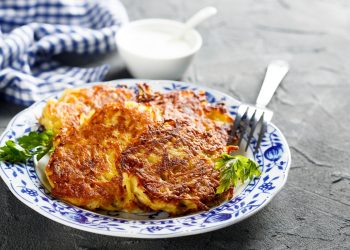 Su tavolo di marmo, piatto bianco con fiori blu con rosti di patate e sotto insalata verde, sul bordo del piatto 1 forchetta e dietro ciotola piccolo con salsa di yogurt e un canovaccio