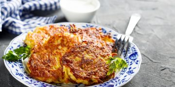 Su tavolo di marmo, piatto bianco con fiori blu con rosti di patate e sotto insalata verde, sul bordo del piatto 1 forchetta e dietro ciotola piccolo con salsa di yogurt e un canovaccio