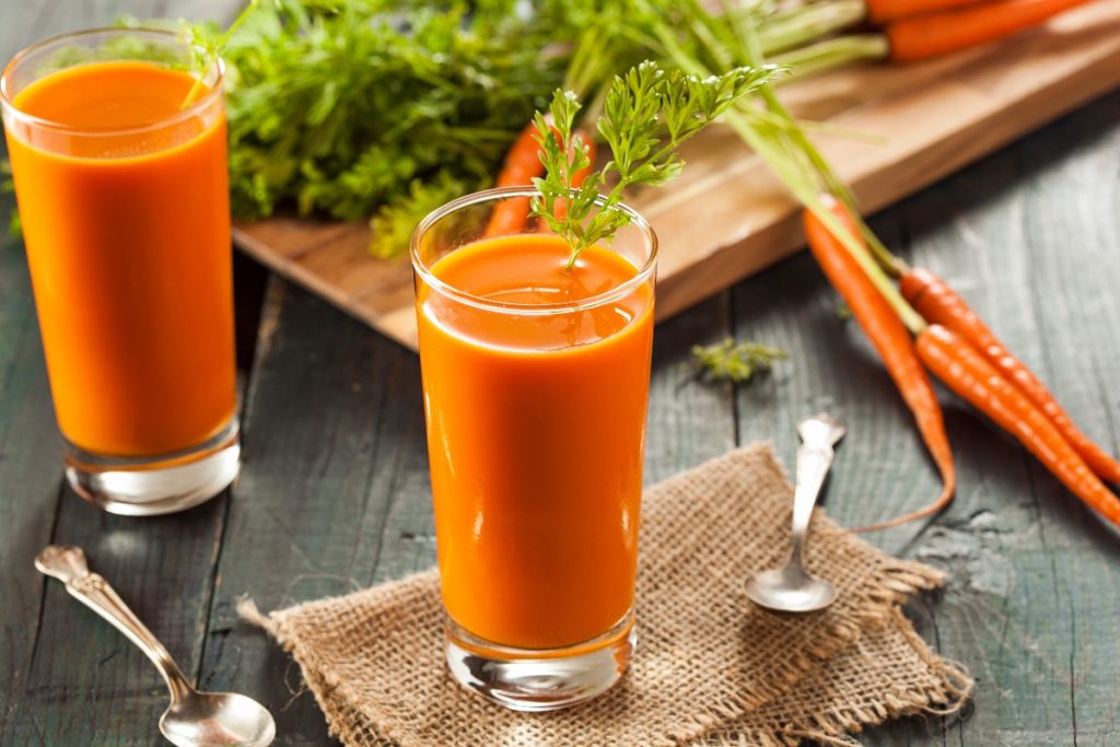 Succo di carota fatto in casa: benefici e ricetta con estrattore e Bimby 3 su tavolo di legno, bicchiere con succo di carota e prezzemolo, dietro tagliere con mazzo di carote