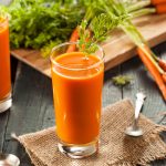 su tavolo di legno, bicchiere con succo di carota e prezzemolo, dietro tagliere con mazzo di carote