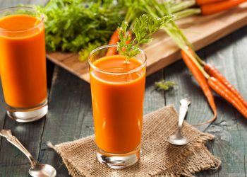 su tavolo di legno, bicchiere con succo di carota e prezzemolo, dietro tagliere con mazzo di carote