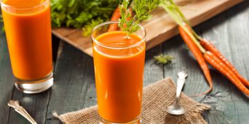 su tavolo di legno, bicchiere con succo di carota e prezzemolo, dietro tagliere con mazzo di carote