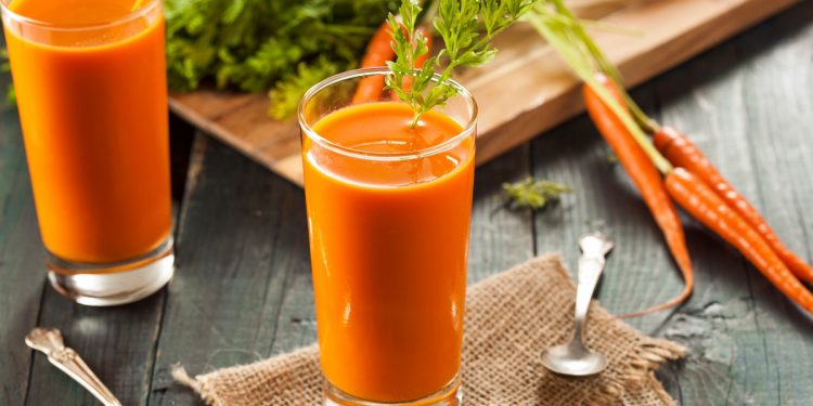 su tavolo di legno, bicchiere con succo di carota e prezzemolo, dietro tagliere con mazzo di carote