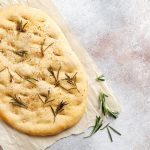 in primo piano su della carta forno una focaccia bianca con sale grosso e rametti di rosmarino