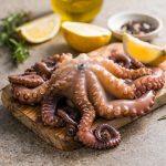 polpo in pentola a pressione: su tavolo tagliere con un polo in pentola a pressione, erbe aromatiche e limone tagliato, dietro una bottiglietta di olio EVO