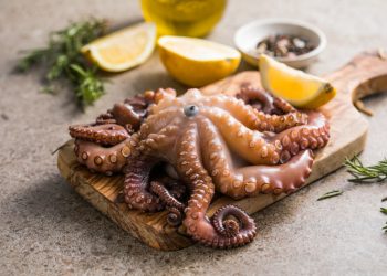polpo in pentola a pressione: su tavolo tagliere con un polo in pentola a pressione, erbe aromatiche e limone tagliato, dietro una bottiglietta di olio EVO