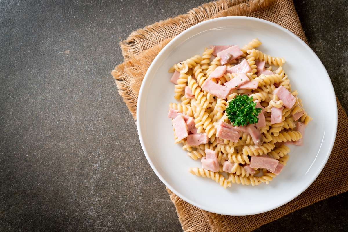 su tavolo di marmo: sotto piatto di paglia con un piatto bianco di pasta con prosciutto cotto e prezzemolo