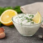 Salsa allo yogurt light: fresca e con poche calorie 1 Su un tavolo da cucina in marmo scuro, ciotola di vetro con la salsa allo yogurt, attorno gli ingredienti limone, erbe aromatiche e aglio.