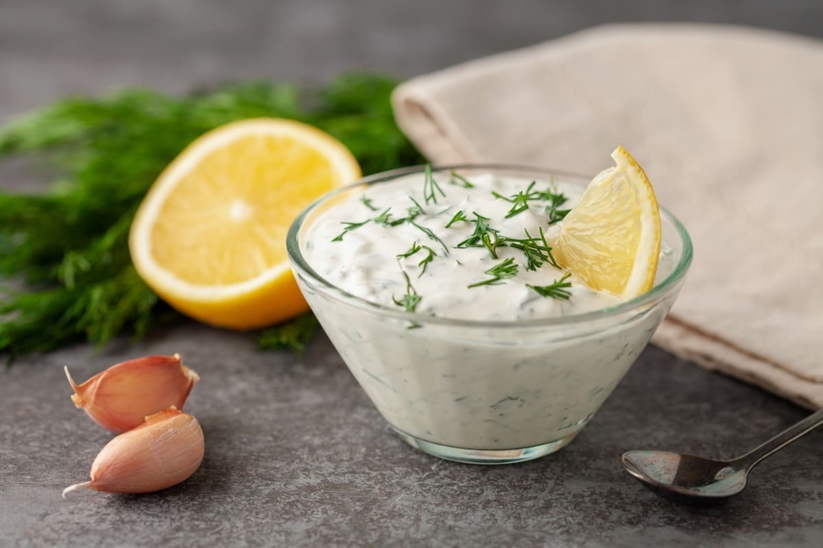Su un tavolo da cucina in marmo scuro, ciotola di vetro con la salsa allo yogurt, attorno gli ingredienti limone, erbe aromatiche e aglio.