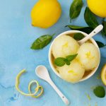 Sorbetto al limone fatto in casa: ricetta facile e cremosa 1 Sorbetto al limone servito in una ciotola bianca con scorza di limone e foglie di menta, su fondo azzurro.