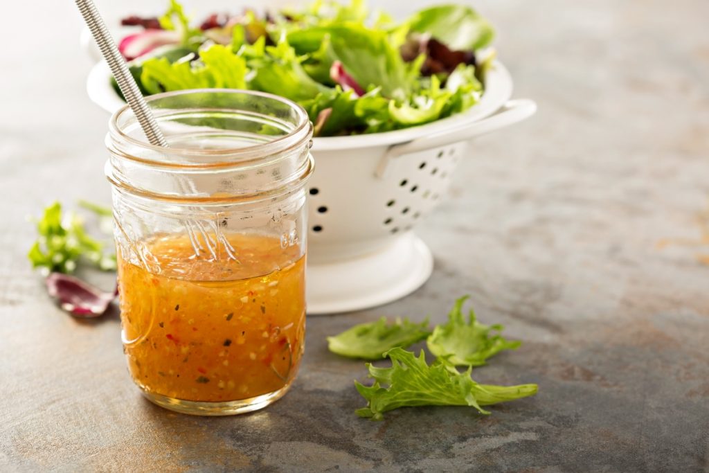 su tavolo ciotola con vinaigrette con frusta, dietro scolapasta in metallo bianco con insalata appena lavata