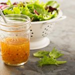 su tavolo ciotola con vinaigrette con frusta, dietro scolapasta in metallo bianco con insalata appena lavata