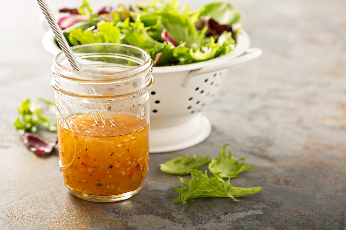 su tavolo ciotola con vinaigrette con frusta, dietro scolapasta in metallo bianco con insalata appena lavata