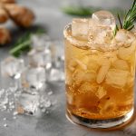 Su tavolo da cucina primo piano di un bicchiere con un ginger ale con ghiaccio e accanto cubetti di ghiaccio, zenzero e erbe aromatiche
