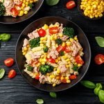 su sfondo nero ciotola nera con pasta fredda con tonno e mais e pomodori. Attorno foglie di basilico e pomodorini tagliati