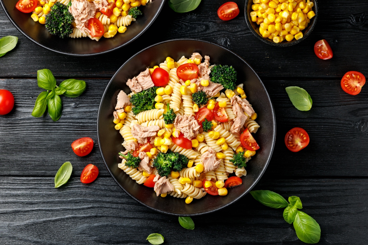 su sfondo nero ciotola nera con pasta fredda con tonno e mais e pomodori. Attorno foglie di basilico e pomodorini tagliati