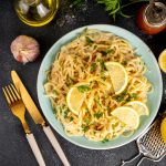 su tavolo di marmo, piatto azzurro con pasta con pesto di limoni e limoni tagliati a fette , accanto forchetto e coltello, limoni e grattugia, olio EVO e erbe aromatiche