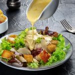in primo piano su un tavolo scuro c'è un piatto di caesar salad che viene condito dalla salsa caesar e crostini croccanti