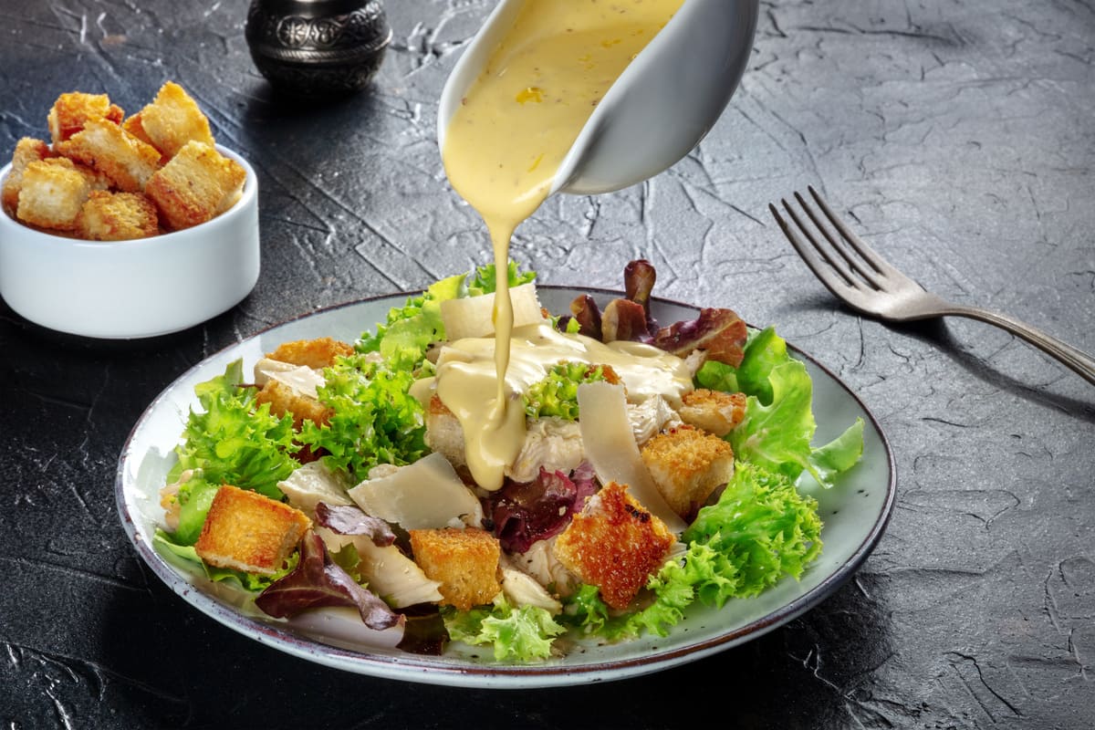 in primo piano su un tavolo scuro c'è un piatto di caesar salad che viene condito dalla salsa caesar e crostini croccanti