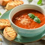 in primo piano un piatto piano color verde acqua con due crostoni di pane su cui poggia una ciotola con della zuppa di pomodoro decorata con una foglia di basilico.