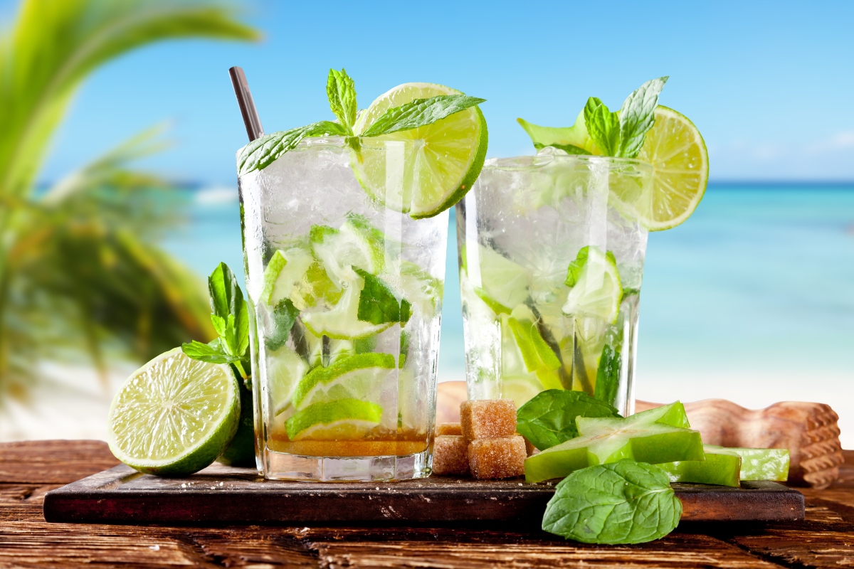 con lo sfondo di una spiaggia tropicale con le palme, tavolo di legno con tagliere e sopra 2 bicchieri con mojito analcolico con lime e foglie di menta e ghiacchio