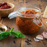 Vasetto di pesto di pomodori secchi con gli ingredienti principali sul tavolo: basilico, aglio, mandorle e parmigiano.