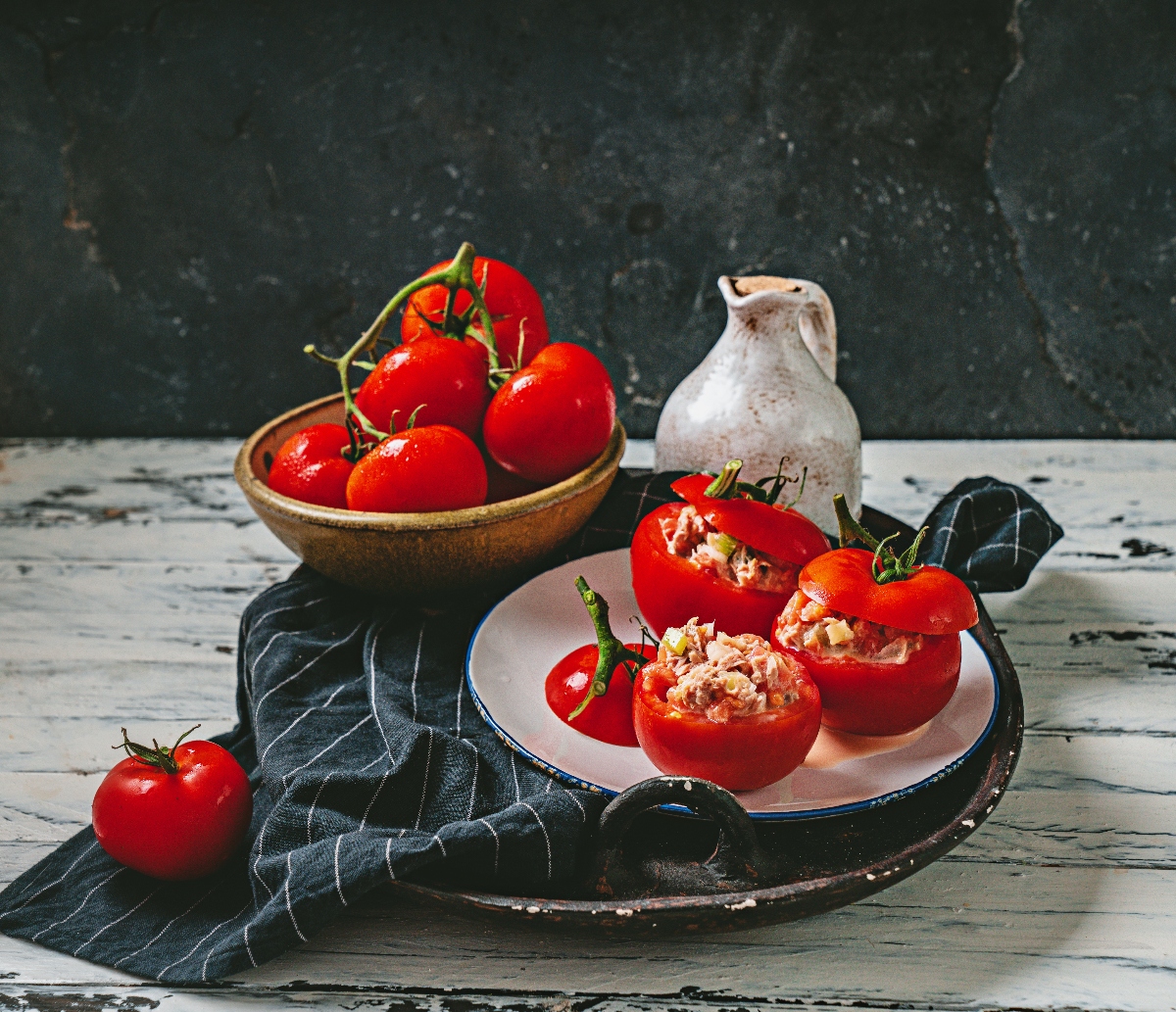 su tavolo di legno bianco piatto con canovaccio nero a righe bianche e un piatto con pomodori ripieni di tonno in scatola, dietro ciottola con pomodori e dietro una brocca d'acqua bianca su sfondo nero