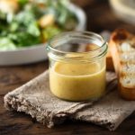 Salsa Caesar fatta in casa: ricetta cremosa e leggera 1 In primo piano su un tavolo di legno, in un barattolino di vetro c'è la salsa caesar fatta in casa e accanto due fette di pane bruscate. Sullo sfondo c'è un piatto di caesar salad.