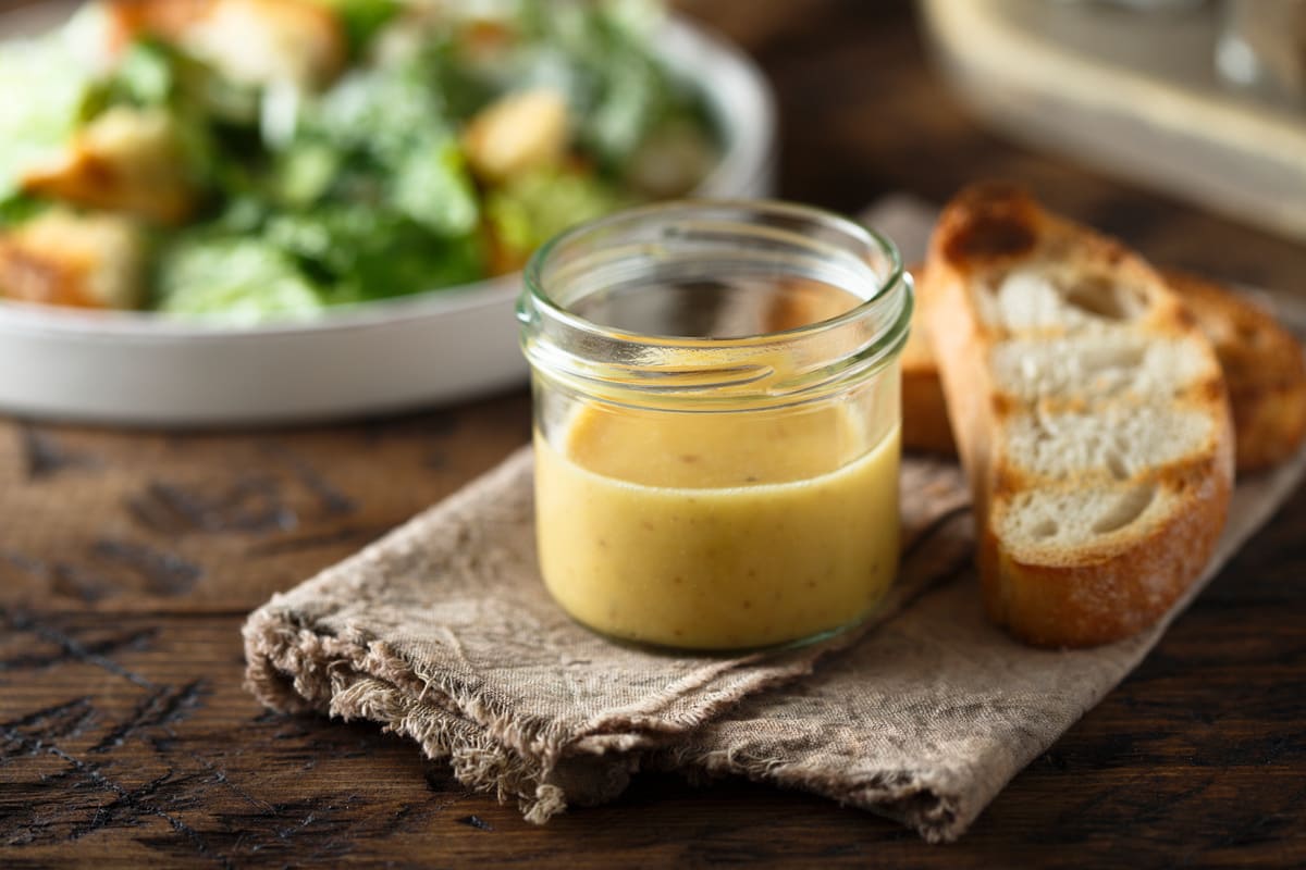 In primo piano su un tavolo di legno, in un barattolino di vetro c'è la salsa caesar fatta in casa e accanto due fette di pane bruscate. Sullo sfondo c'è un piatto di caesar salad.