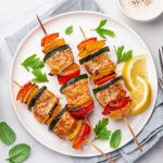 Spiedini di pesce al forno morbidi e ben dorati 1 In primo piano su un tavolo bianco, c'รจ un piatto fondo con degli spiedini di pesce e verdure. Sul piatto ci sono due spicchi di limone e delle erbette aromatiche.