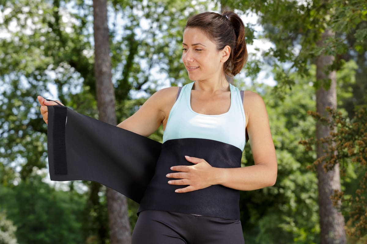 Una donna in tenuta sportiva applica una fascia addominale dimagrante nera intorno alla vita mentre si prepara all’attività fisica all’aperto. L’immagine rappresenta l’utilizzo delle fasce addominali dimagranti come supporto per l’allenamento e il controllo della sudorazione.