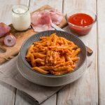 su tavolo di legno piatto fondo con penne al baffo e accanto tagliere con gli ingredienti cipolla, panna, prosciutto cotto e sugo di pomodori