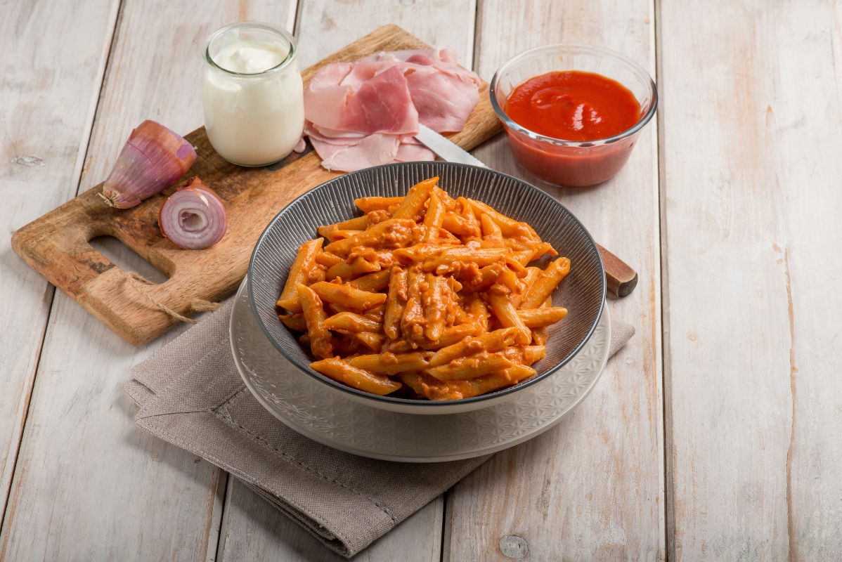 su tavolo di legno piatto fondo con penne al baffo e accanto tagliere con gli ingredienti cipolla, panna, prosciutto cotto e sugo di pomodori