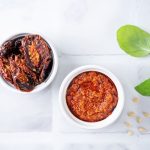 Pesto di pomodori secchi: ricetta facile e versatile 1 Pesto di pomodori secchi in una ciotola bianca, accanto a pomodori secchi, foglie di basilico e pinoli su sfondo chiaro.